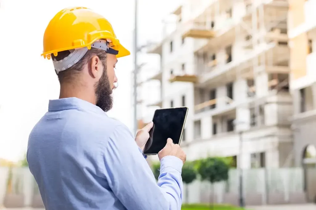 Businessperson wearing hardhat holding tablet standing in front of apartment
buildings.