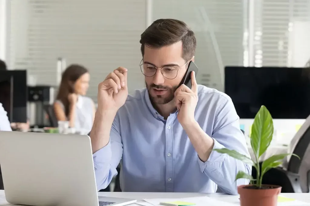 A person in a dress shirt talking on a cell phone in an office with a coworker behind them. 