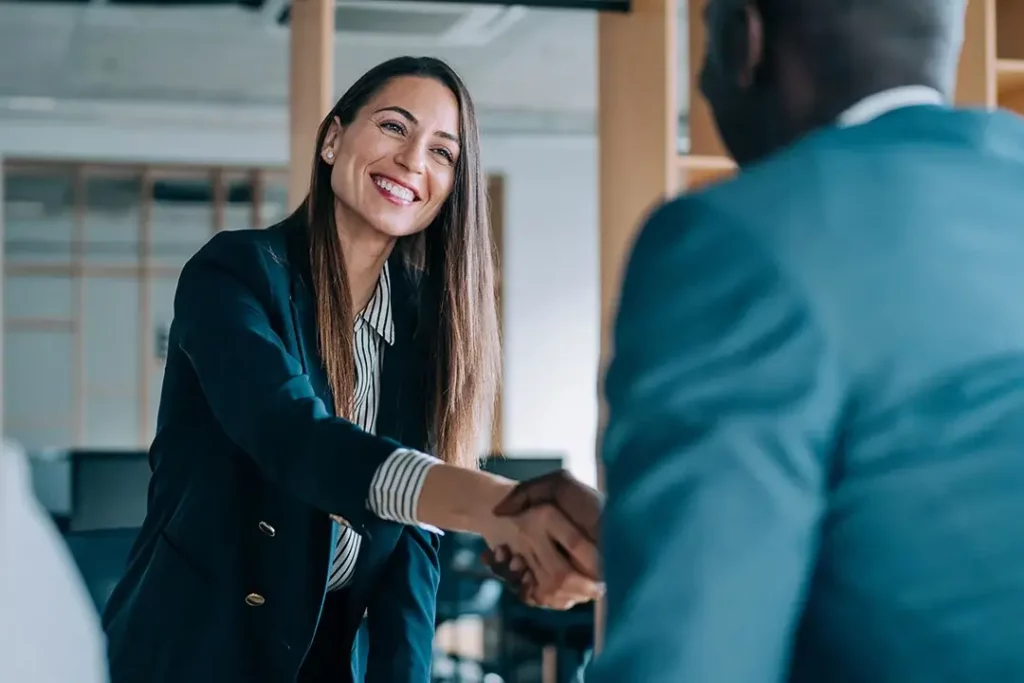 A smiling businessperson shaking hands with someone in a suit in a building.
