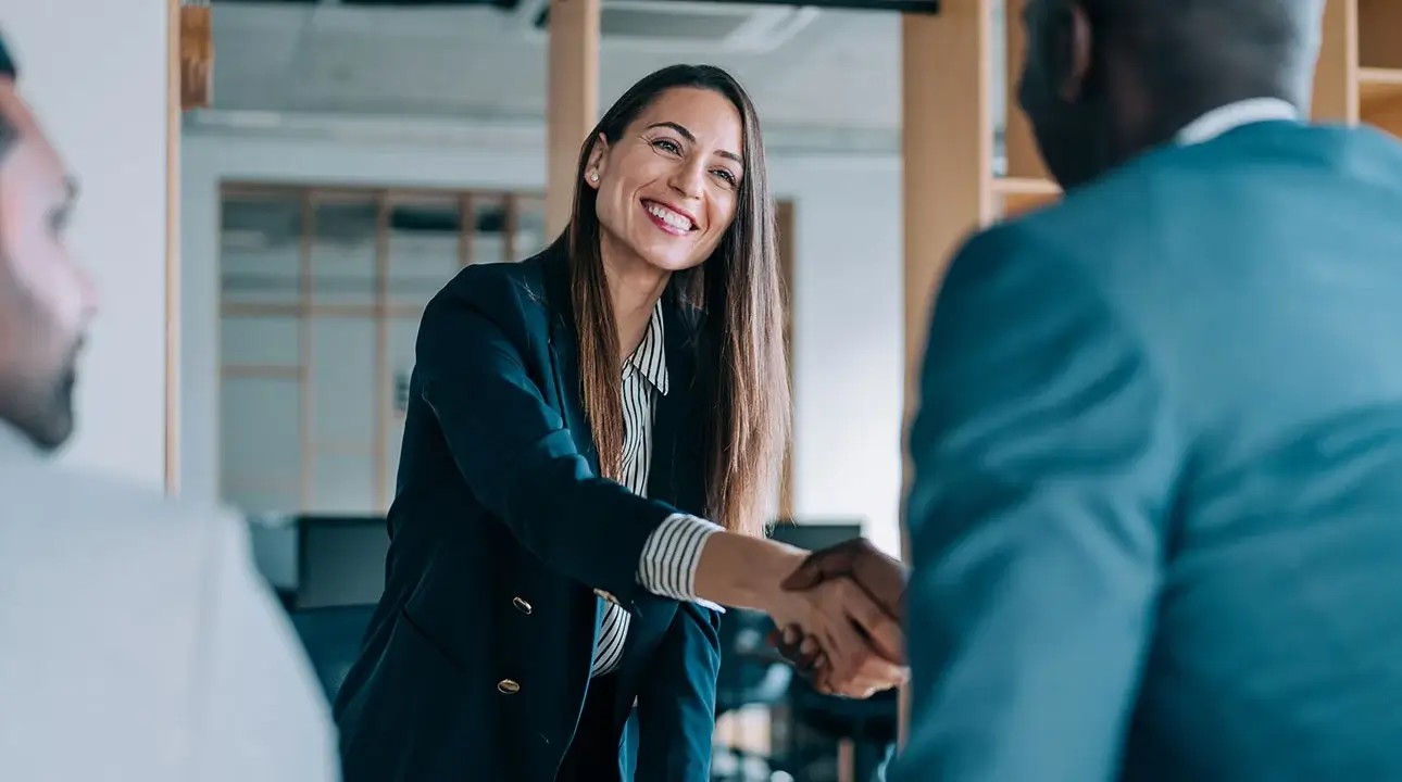 A smiling businessperson shaking hands with someone in a suit in a building.