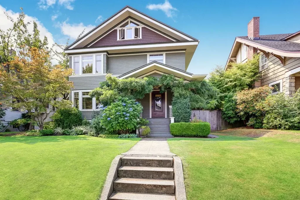 A view looking up at a gray and purple house with shrubs in the front and stone steps.