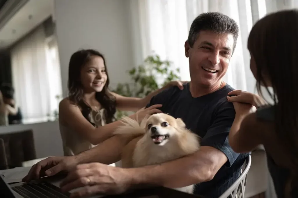 A dad holding the family dog while working on laptop, with two daughters laughing with him.