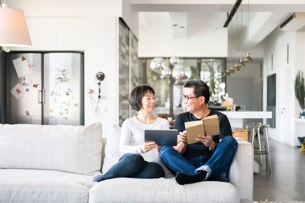 A middle-aged couple, with one holding tablet and one holding a book, sit on a light cough in a bright house.