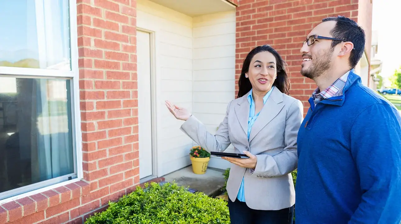 Businessperson holding tablet and gesturing towards house while talking with consumer.