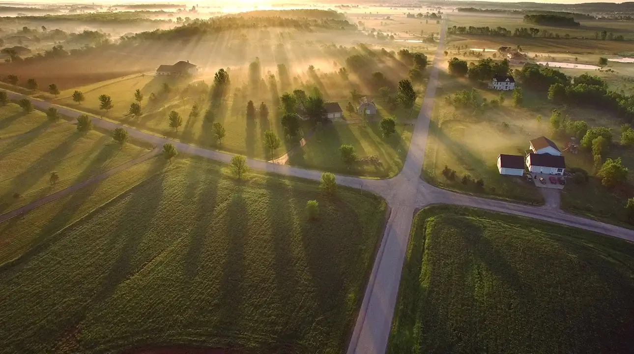 An overview of a crossroads at sunset surrounded by green fields, trees and one house.