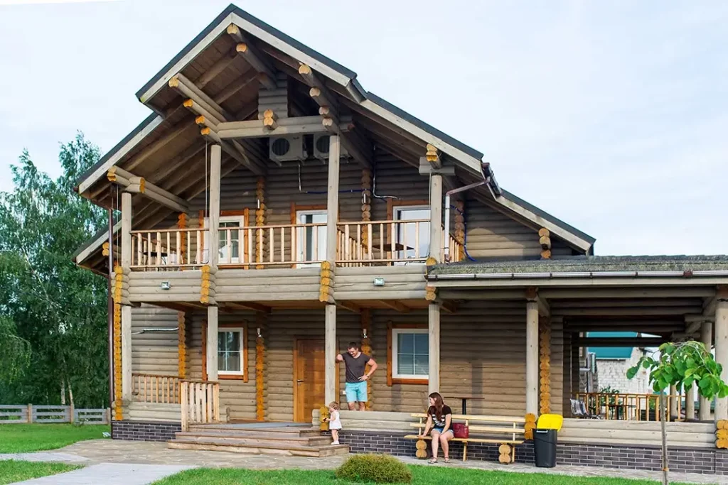 A two-story log cabin house in the summer with two adults and a child in front. 