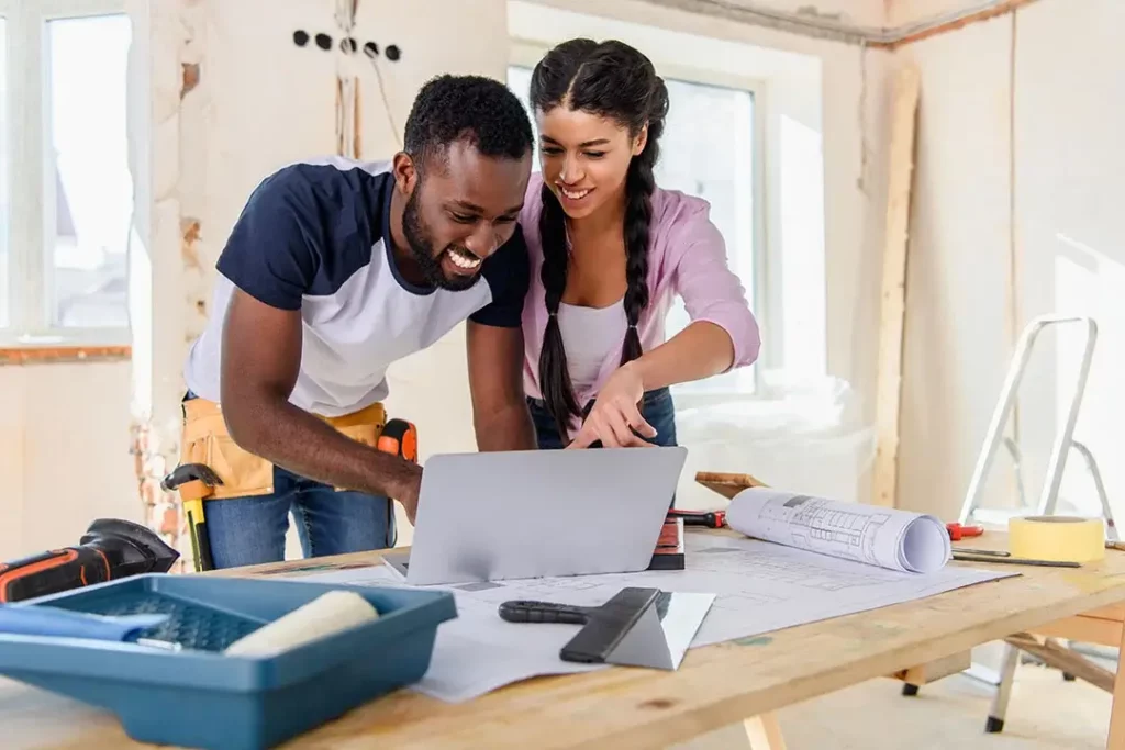 Two smiling people inside a house being renovated looking at a computer. One person is wearing a tool belt.