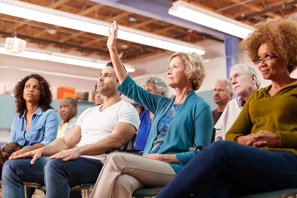 A group of people sitting in a meeting listening, with one person raising their hand.