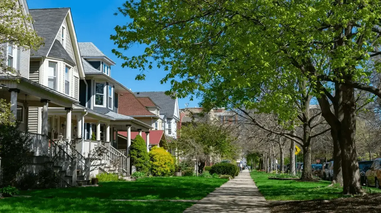 A view down a neighborhood sidewalk with houses on the left and trees on the right.